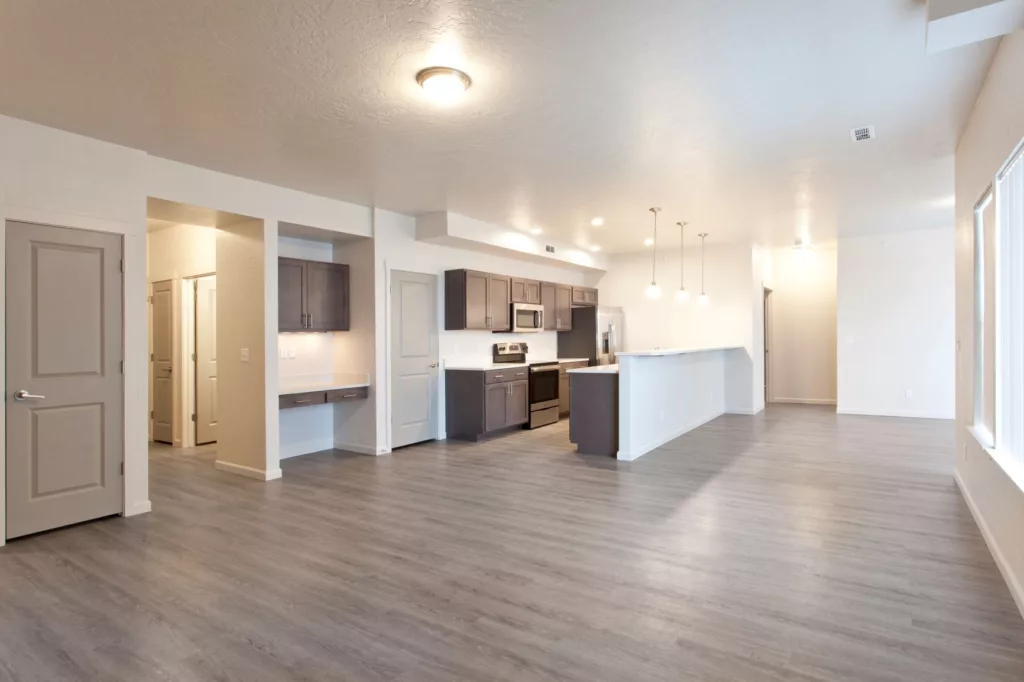 Open-plan kitchen and living area with light gray wood flooring, dark cabinetry, stainless steel appliances, pendant lights, and ample natural light from large windows.