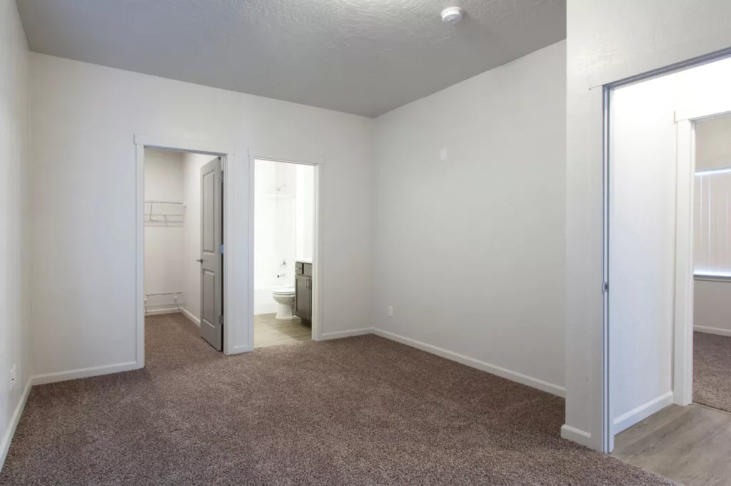 An empty room with beige carpet, two open doorways, one leading to a bathroom with visible toilet and sink, and another leading to a closet with shelves.