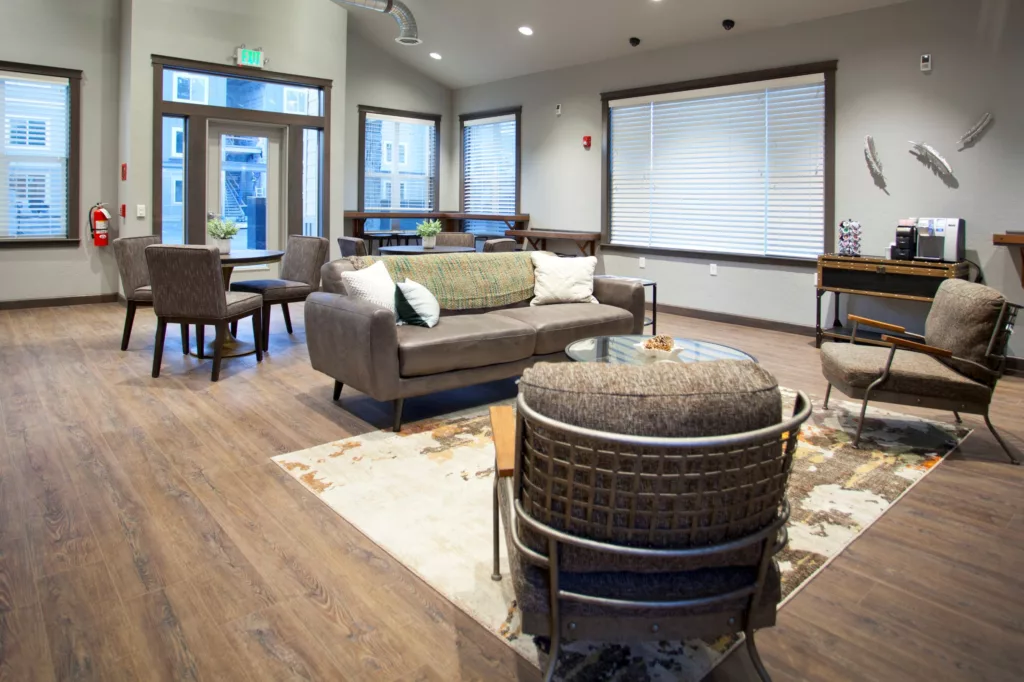A modern living room with wooden flooring, featuring a gray sofa with pillows, a glass coffee table, and cushioned chairs. Large windows with blinds line the walls, and a small table with chairs is in the corner. Decorative objects and a coffee machine are on a side table.