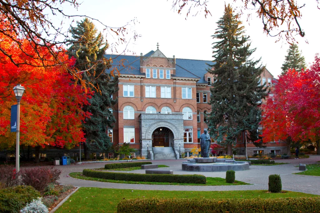 A historic building with a brick facade stands amidst vibrant autumn foliage, featuring a statue and circular pathways in the foreground.