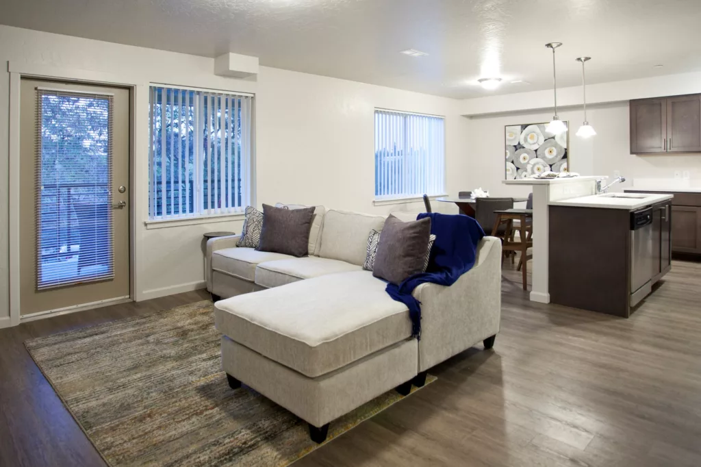 Modern open-plan living room and kitchen with a light gray sectional sofa, dark wood cabinets, and a dining area, featuring a glass door and large windows with vertical blinds.