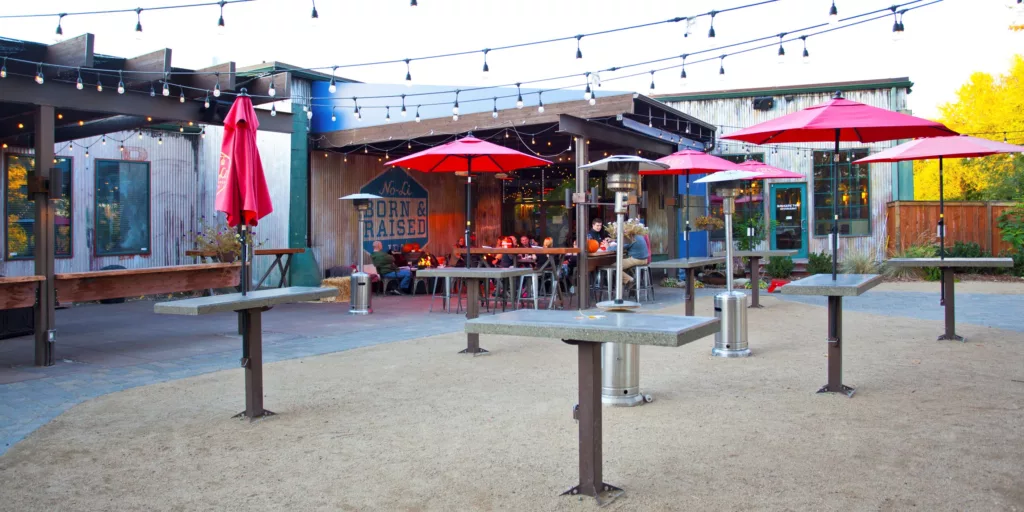 Outdoor seating area of a restaurant with red umbrellas over tables, string lights above, and a group of people sitting at a table in the background. The establishment has a rustic look with corrugated metal siding and a sign reading 'Born & Raised.'