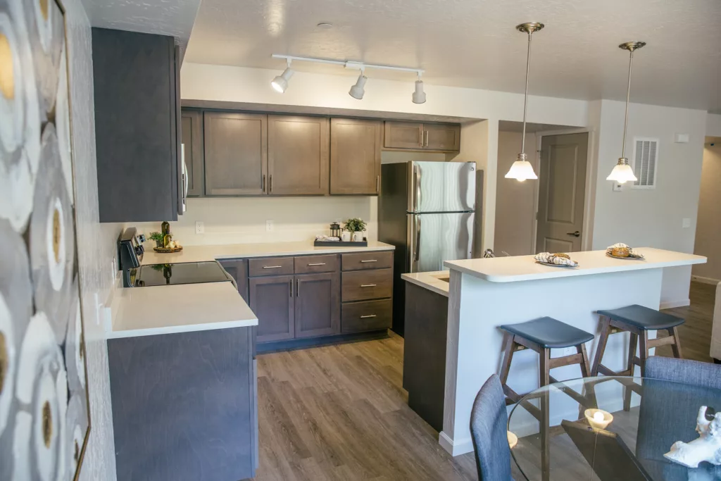 Modern kitchen with dark wood cabinets, stainless steel appliances, and a breakfast bar with two stools. The kitchen features track lighting and pendant lights above the bar. A dining area with a glass table is visible in the foreground.