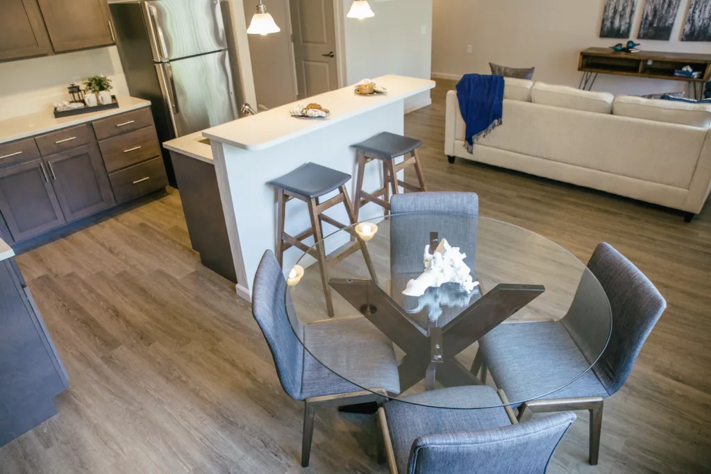 A modern kitchen and dining area featuring a round glass table with four gray upholstered chairs. There is a breakfast bar with two bar stools and snacks on the counter. The kitchen has dark wood cabinets and a stainless steel refrigerator. In the background, a beige sofa with a blue throw is visible in the living area.